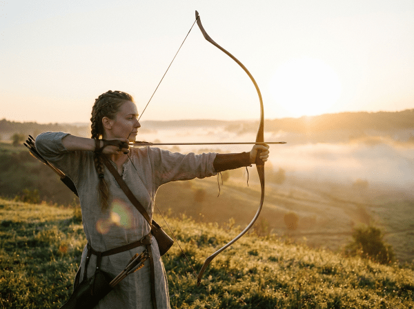 Woman in medieval attire aims a bow across a misty valley at sunrise.