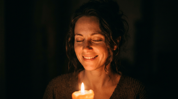 Smiling woman with closed eyes holding a lit candle in darkness
